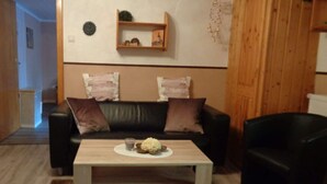 A living room with a black leather sofa, two cushions, a wooden coffee table with a glass and a bowl, and a wooden wall shelf with decorative items. - Fantastic Apartment in Reinhardshagen (Reinhardshagen)