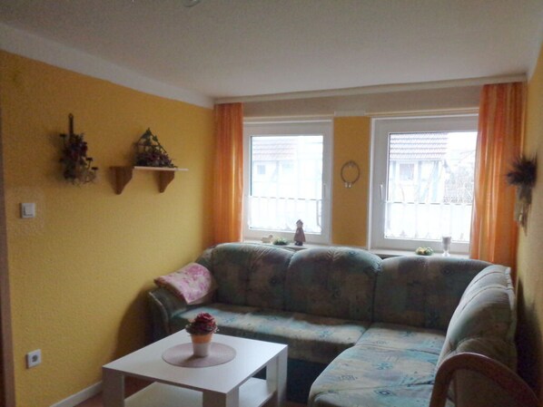 Ein Wohnzimmer mit einem Ecksofa, einem kleinen Tisch und zwei mit Vorhängen versehenen Fenstern.