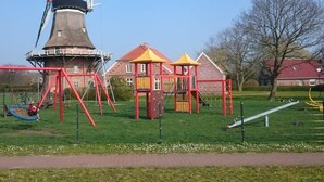 A playground with swings and climbing structures, a windmill, and a brick building in the background.