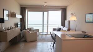 A modern hotel room with a sofa, a dining table, a flat-screen TV, a lamp, and a view of the sea through the sliding doors. - Apartment in Nieuwpoort Near the Beach (Nieuwpoort)