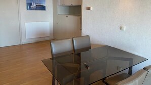A modern dining area with a glass table and leather chairs, a white radiator, a framed picture of a sailboat, and a kitchenette with cabinets. - Apartment in Nieuwpoort Near Sandy Beaches (Nieuwpoort)