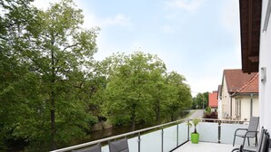A balcony with a table and chairs overlooking a river, surrounded by greenery and buildings. - Lovely Apartment in Himmelkron (Himmelkron)