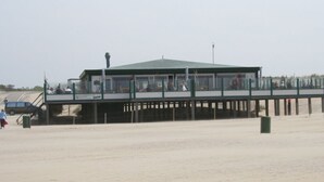 Un muelle frente a la playa con una zona de estar cubierta y un pequeño edificio, probablemente un café o restaurante, con vistas a la playa.