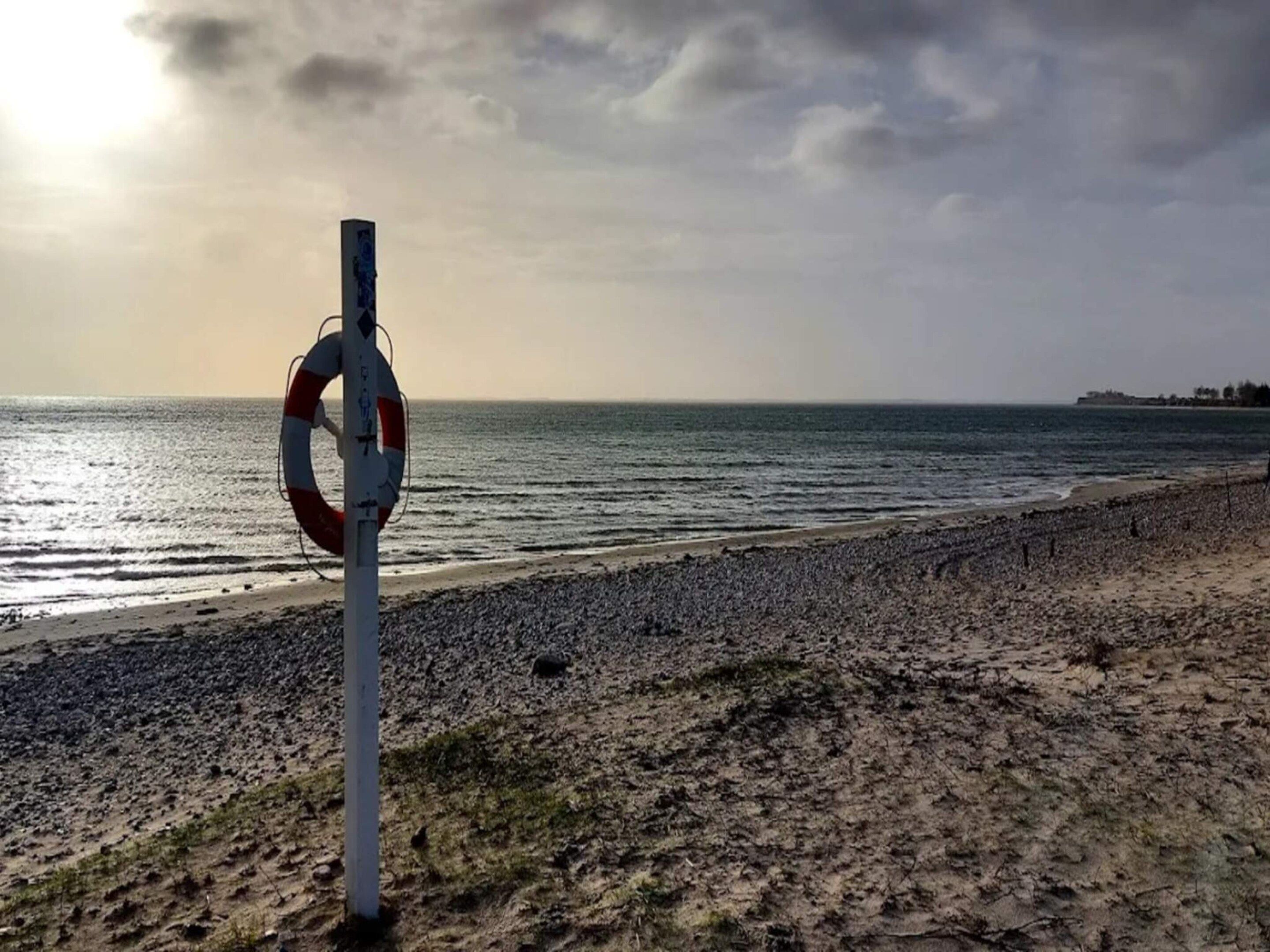 Une bouée de sauvetage est fixée à un poteau sur une plage de galets, avec l’océan en arrière-plan.