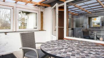 Un patio cubierto con una mesa redonda, una silla de mimbre, ventanas amplias y vistas al exterior.