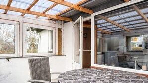 Un patio techado con una mesa redonda y una silla de mimbre, una puerta de vidrio que da a un espacio interior y vistas al exterior.
