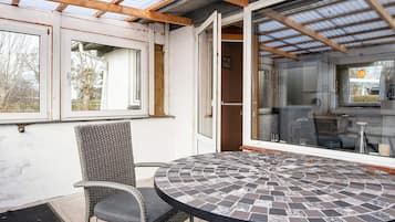 A covered patio area with a round table and a wicker chair, a glass door leading to an interior space, and a view of the outside.