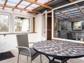 Un patio techado con una mesa redonda y una silla de mimbre, una puerta de vidrio que da a un espacio interior y vistas al exterior.