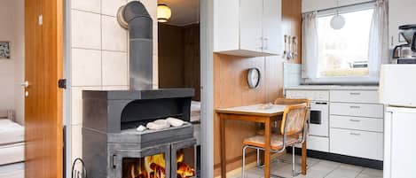 Een kleine keuken met een houtkachel, een eethoek met een tafel en stoelen, en een raam met gordijnen.
