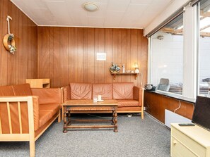 A room with a wooden panel wall, a brown leather sofa, a wooden coffee table, a small desk with a computer, and a window with a view of the outside. - 3 Person Holiday Home in Sydals (Sydals)