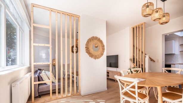 A modern living room with a wooden partition, a round woven mirror, a dining table with white chairs, and a TV on the wall. - Garden Villa Steps From Beaches (Koksijde)