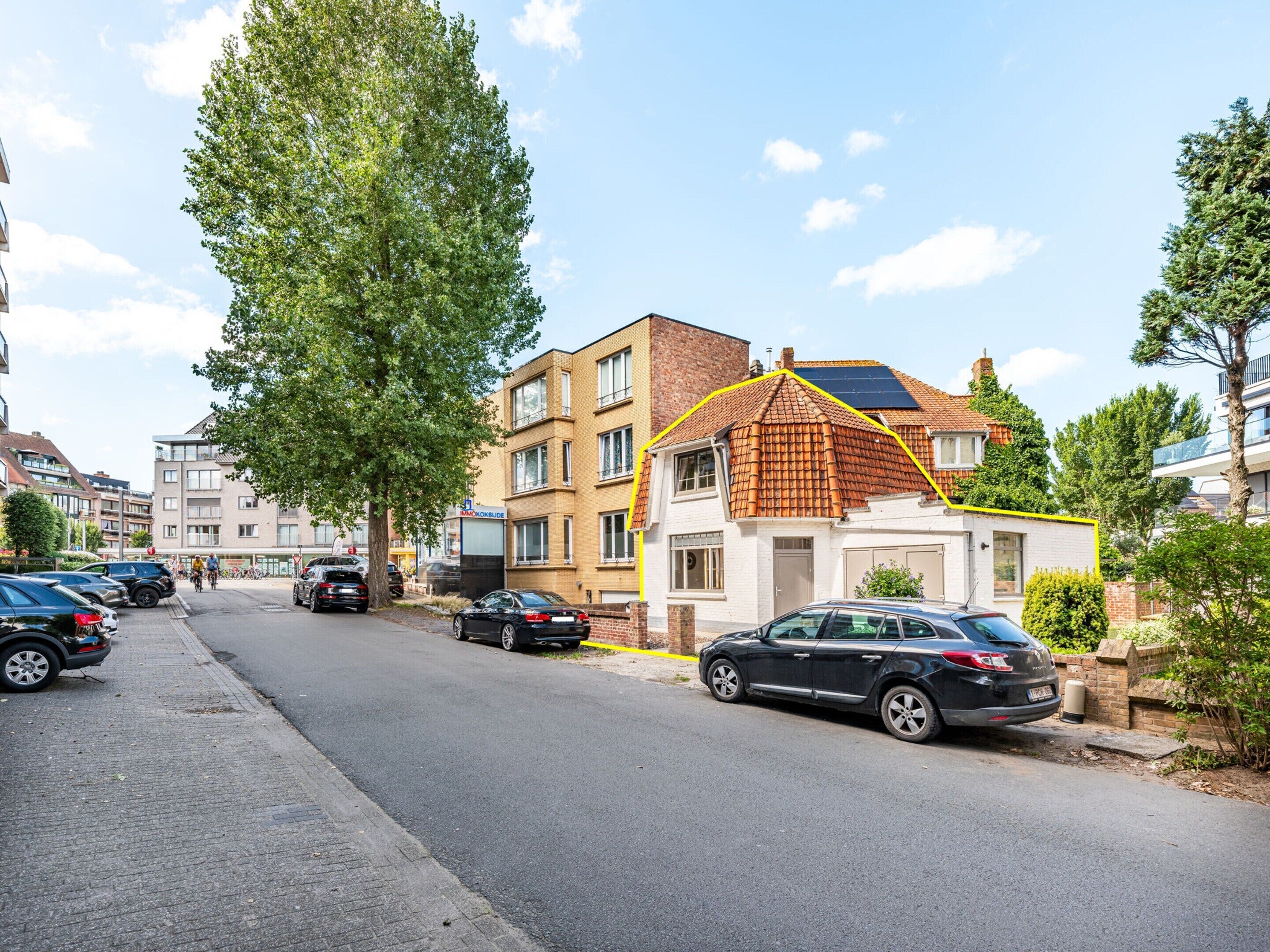 A residential street with parked cars, a large tree, and multi-story buildings.