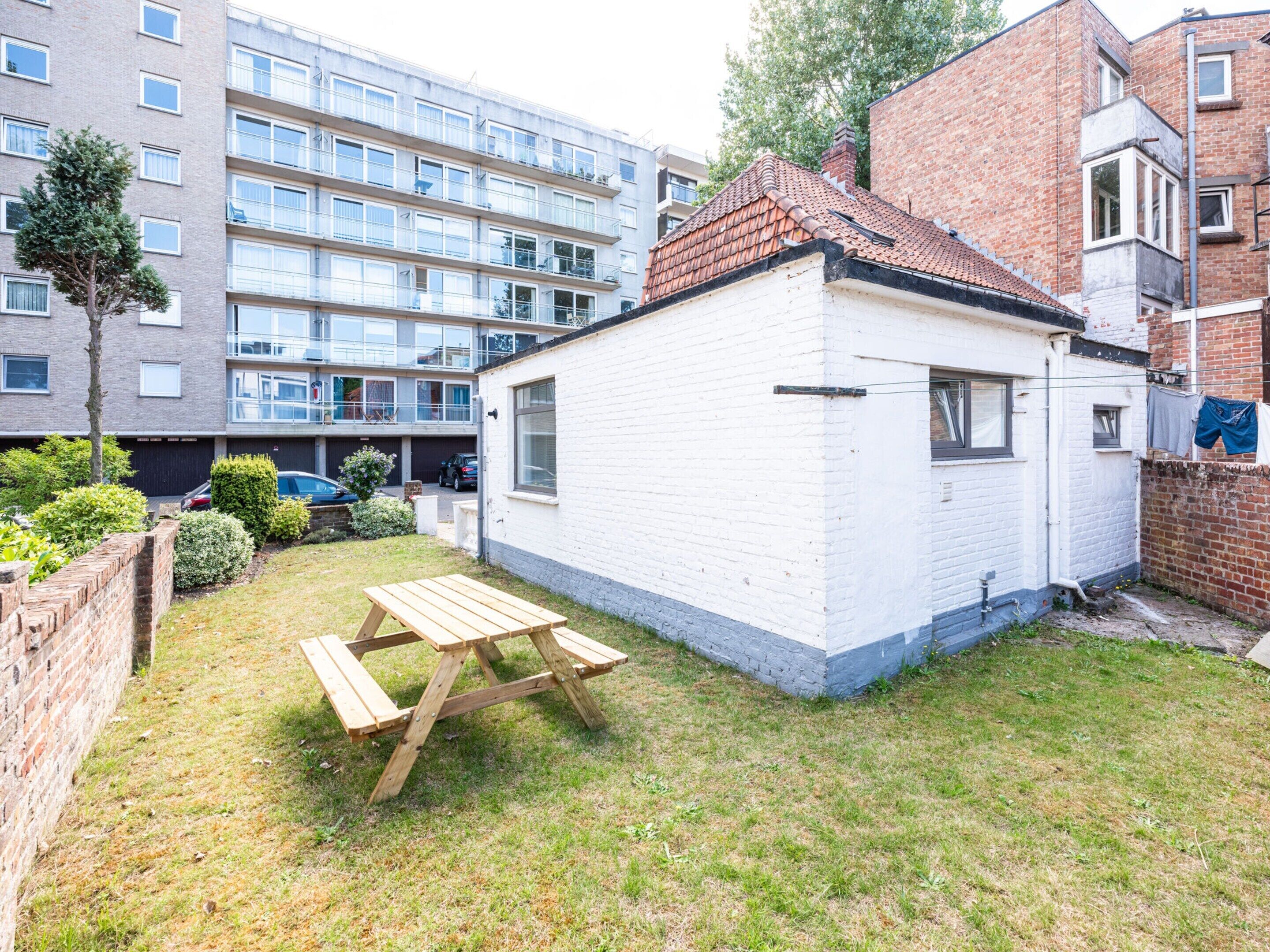 A small white house with a tiled roof, a wooden picnic table, and a brick wall.