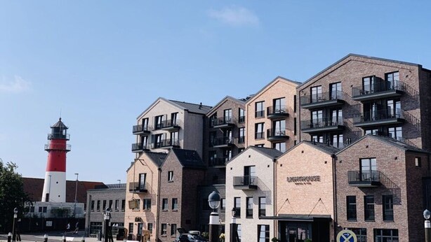 A row of modern brick buildings with balconies, a lighthouse in the background, and a clear blue sky. - "norderney" in the Achter de Waterkant House (Büsum)