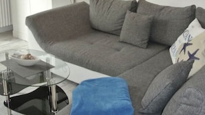 A living room with a grey sofa, a glass coffee table, and a blue cushion. There is a white cabinet with drawers and a small plant on top. A painting of a beach is on the wall. - "norderney" in the Achter de Waterkant House (Büsum)