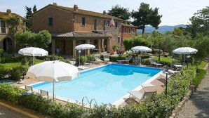A pool area with lounge chairs and umbrellas, surrounded by greenery and a building with a tiled roof. - Apartment in Bolgheri Near Tuscan Beaches (Castagneto Carducci)