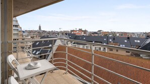 Un balcón con mesa y sillas, con vista a un paisaje urbano que incluye varios edificios y una torre distante.