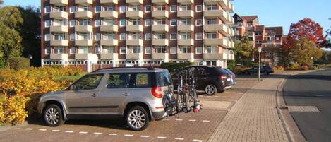 A multi-story residential building with balconies, a paved sidewalk, and parked cars.