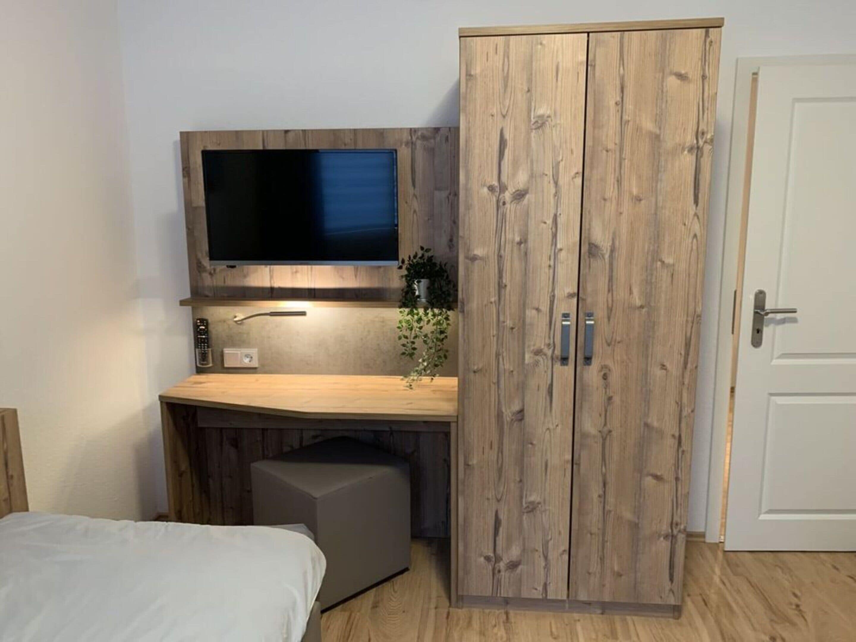 A bedroom with a bed, a wooden wardrobe, a TV mounted on the wall, and a small desk area.