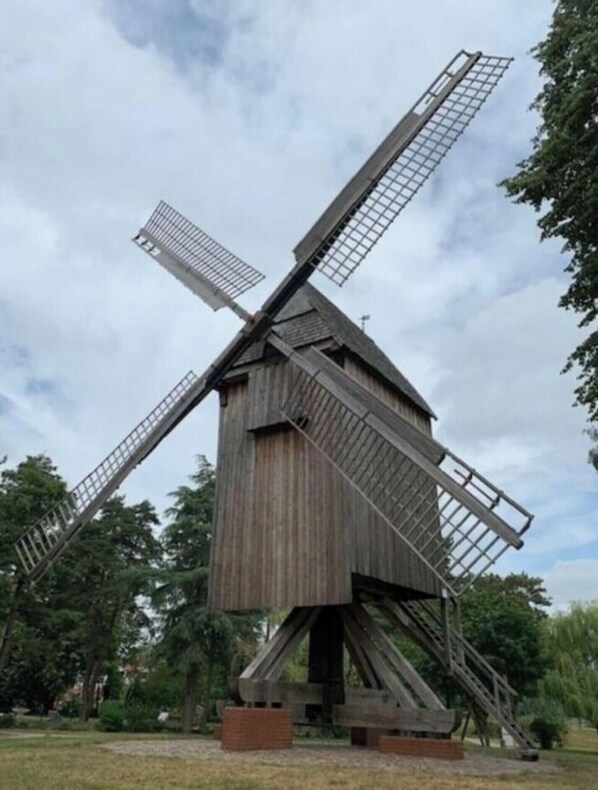 Eine große, hölzerne Windmühle mit Gitterstruktur, eingebettet in eine grüne Wiese mit Bäumen im Hintergrund.