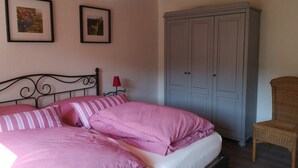 A bedroom with a bed, two framed pictures on the wall, a wardrobe, and a wicker chair. - Holiday Apartment at the Linden Tree (Dernbach)