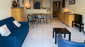 En moderne stue med blå sofa, spisebord med stole, et køkkenområde og et fjernsyn på et stativ.