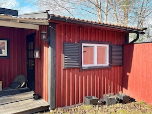 A red wooden building with a window and shutters, a door, and a small porch area with a fan and potted plants.