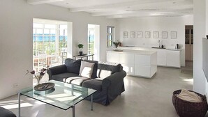 A modern living room with a glass coffee table, a sofa, and a kitchen area with a white island and framed pictures on the wall. - 5 Star Holiday Home in Assens (Assens)
