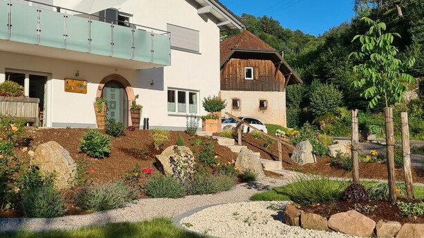 A modern two-story house with a balcony, a wooden barn-like structure, and a well-maintained garden with rocks and plants. - Lovely Apartment in Baden-wurttemberg (Oberkirch)