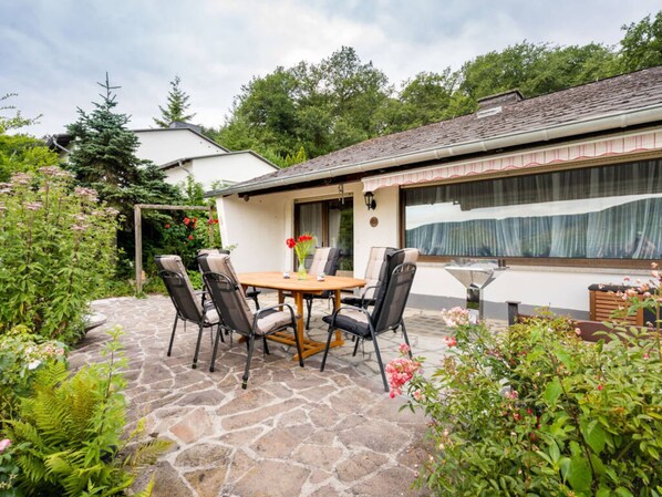 A patio with a stone table and chairs, surrounded by lush greenery and a house with a large window. - Holiday Home Villa Bauer (Traben-Trarbach)