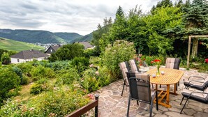A patio with a stone table and chairs, surrounded by lush greenery and a view of a village in the background. - Holiday Home Villa Bauer (Traben-Trarbach)