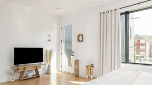 A modern bedroom with a large bed, a TV on a wooden stand, a door with glass panels, and a view of buildings outside.