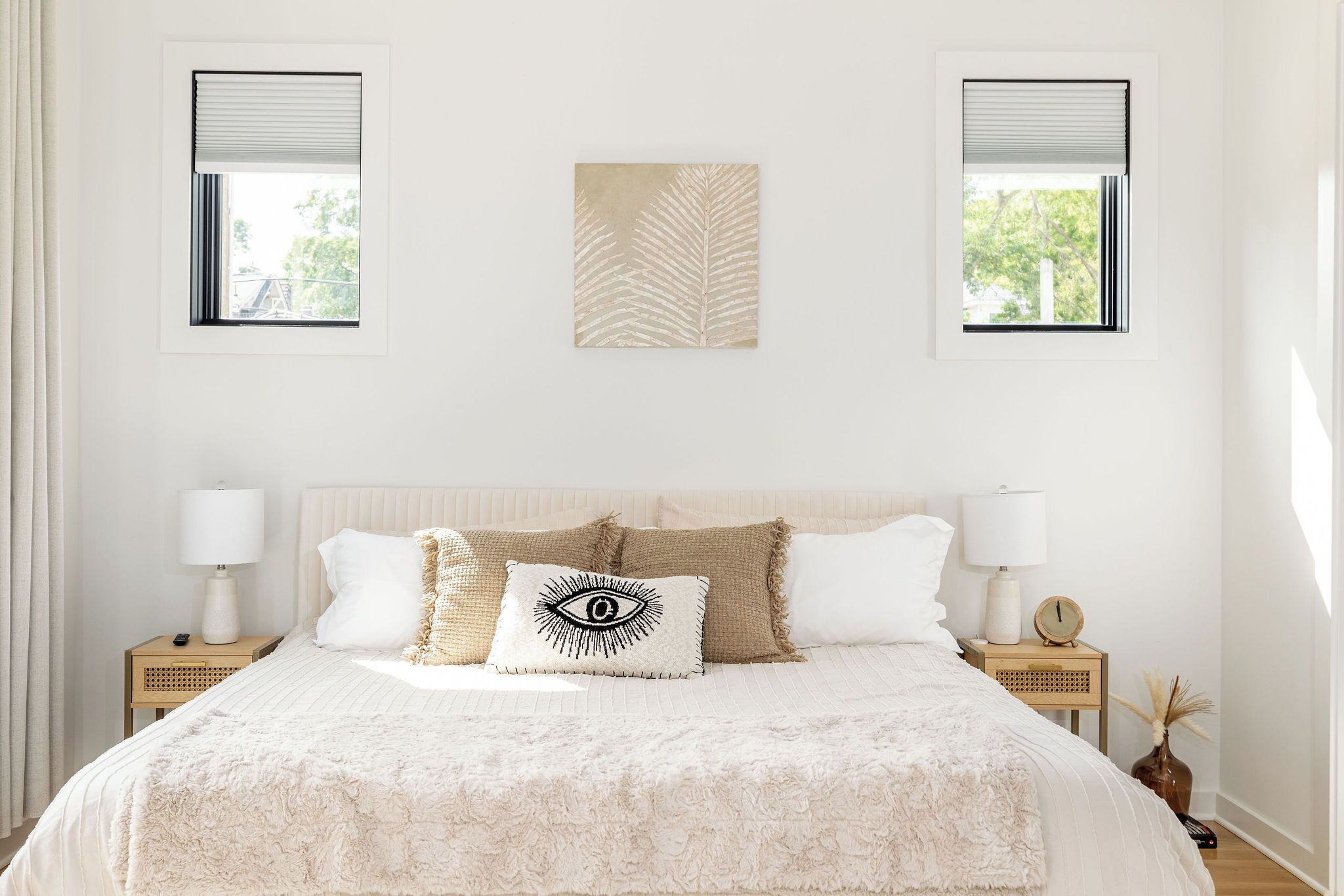A bedroom with a large bed, two bedside tables, a nightstand, a wall-mounted artwork, and two windows with blinds.