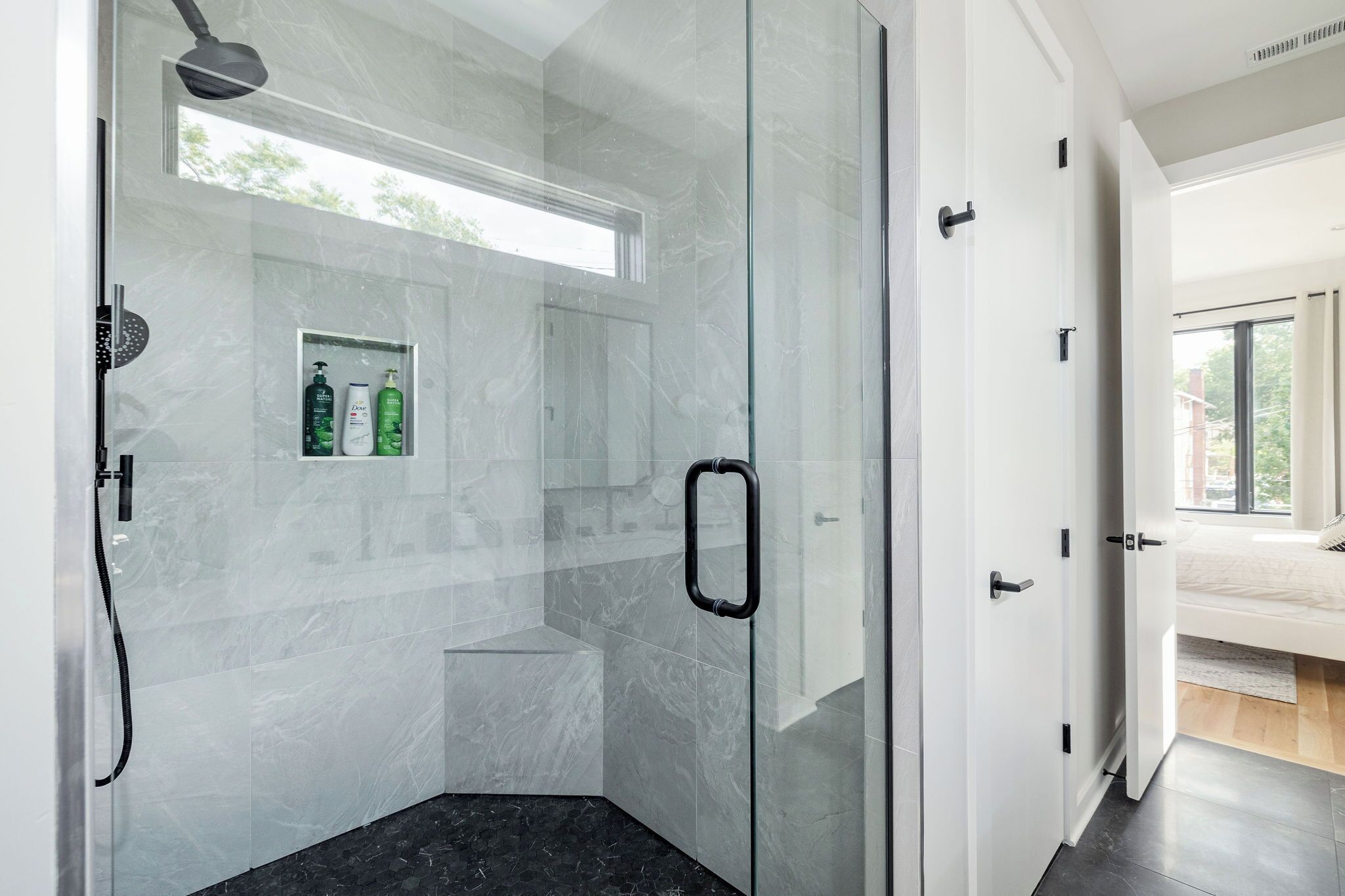 Une salle de bain moderne dotée d’une douche à parois de verre, d’une étagère intégrée pour les articles de toilette et offrant une vue sur la chambre adjacente.
