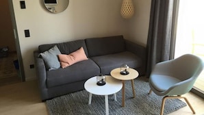 A modern living room with a grey sofa, two small round tables, a blue armchair, a round mirror on the wall, and a hanging light fixture. - 8 in het Huis Meerloft (Norderney)