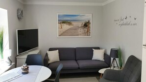 A modern living room with a grey sofa, a flat-screen TV, a dining table with a striped tablecloth, and a beach-themed wall art. - Romantic Apartment Near Seabeach (Büsum)