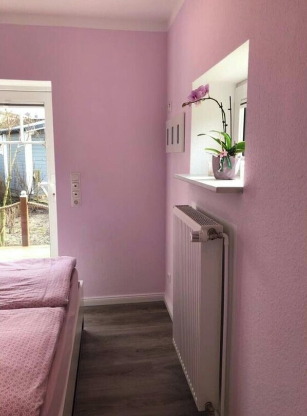 A narrow hallway with a radiator, a bed with pink bedding, and a shelf with potted plants.