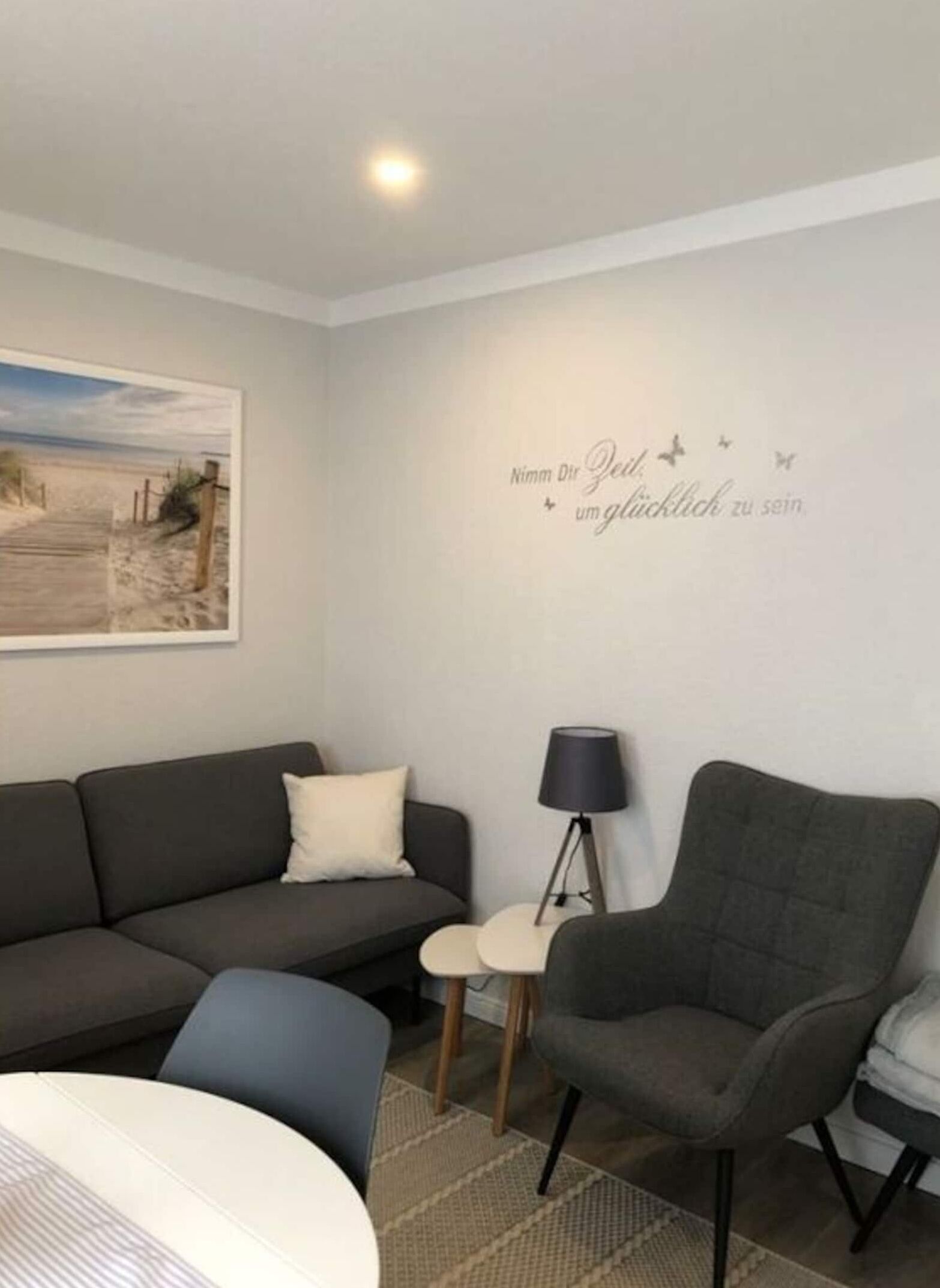 Ein modernes Wohnzimmer mit einem grauen Sofa, einem kleinen Sessel und einem Beistelltisch mit einer Lampe. An der Wand ist eine Strandansicht zu sehen.