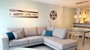 A modern living room with a grey sectional sofa, a glass coffee table, a wall clock, and a dining area with a white table and chairs. - Apartment in Nieuwpoort Near the Beach (Nieuwpoort)