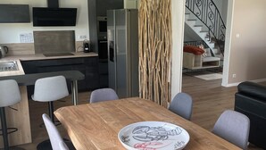 A modern kitchen with a built-in oven, a refrigerator, and a dining area with a wooden table and grey chairs. - Renovated house near beaches and golf (Bénodet)