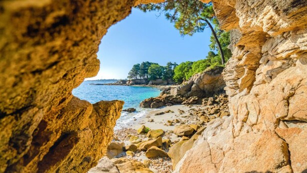 A coastal view through a rocky archway with a beach and trees in the background. - Renovated house near beaches and golf (Bénodet)