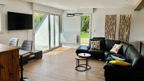 A modern living room with a black leather sofa, a wooden dining table, and a flat-screen TV mounted on the wall. - Renovated house near beaches and golf (Bénodet)