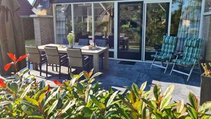 Une terrasse extérieure avec une table et des chaises, une porte-fenêtre coulissante donnant sur un jardin, et un petit coin salon avec des fauteuils.