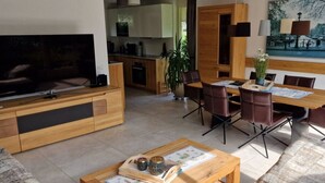 Un salon moderne avec un téléviseur, des meubles en bois, un coin repas avec des chaises et une cuisine en arrière-plan.
