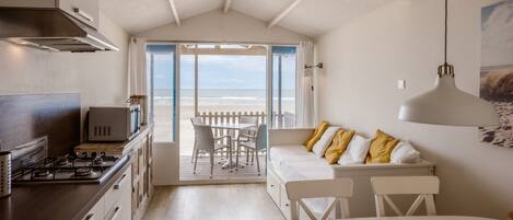 A kitchen with a stove, microwave, and cabinets. There is a dining area with a table and chairs, and a sofa with cushions. A large window offers a view of the beach.