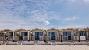 A row of beach huts with blue doors and white railings, arranged on a sandy beach under a partly cloudy sky. - Holiday Home in Wijk aan Zee by the Beach (Wijk aan Zee)