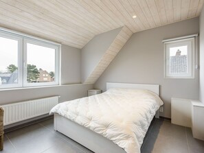 Une chambre avec un plafond en pente, une grande fenêtre, un lit recouvert d’une couette blanche, une table de chevet et un radiateur.