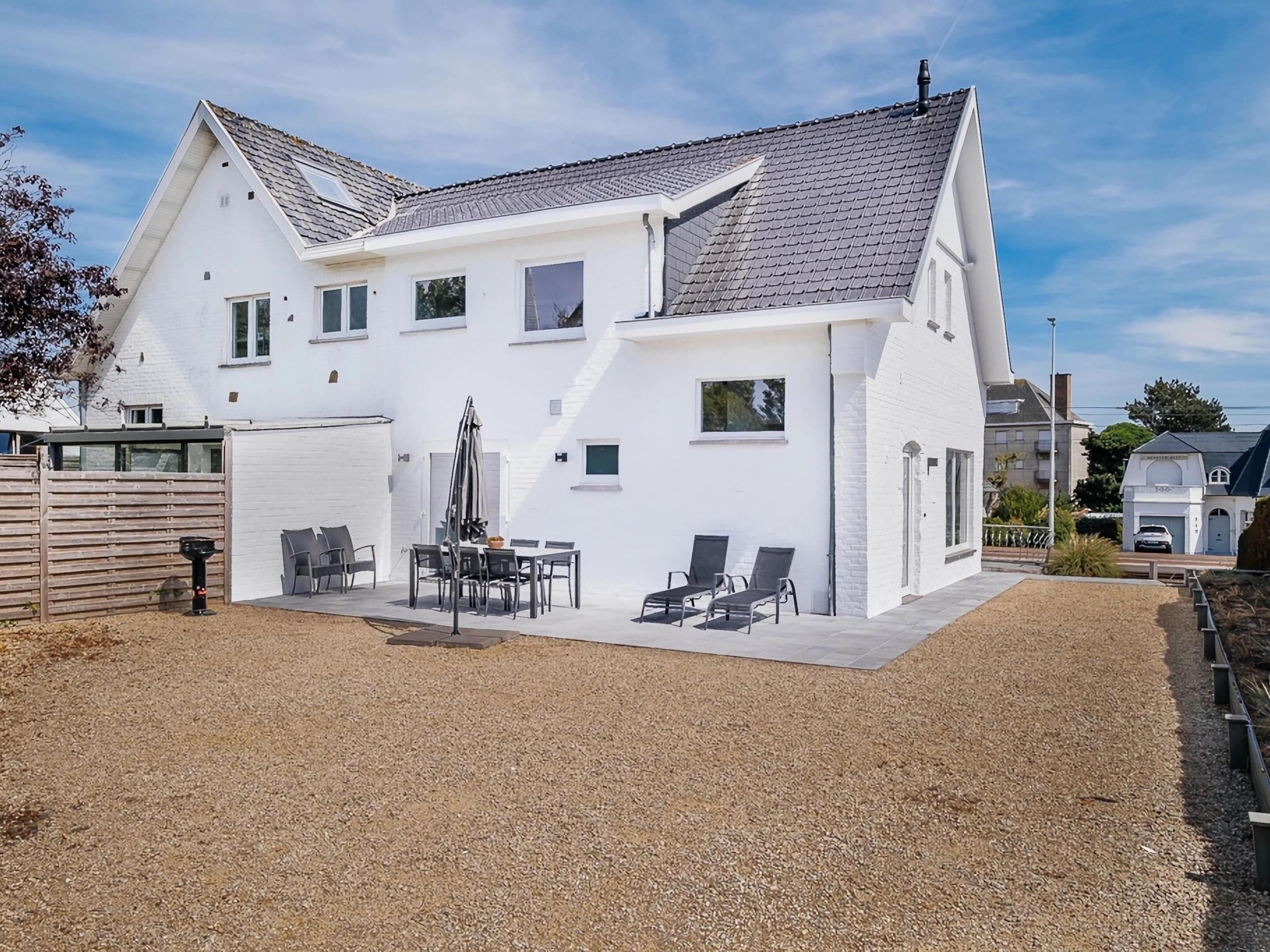 Ein weißes, zweistöckiges Haus mit geneigtem Dach, einer Terrasse mit Gartenmöbeln und einem Schotterweg.