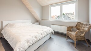 Ein Schlafzimmer mit schrägen Wänden, einem großen Bett, einem Korbstuhl und einem Fenster mit Blick auf Gebäude.