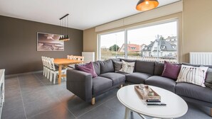 Ein modernes Wohnzimmer mit einer grauen Eckcouch, einem runden weißen Couchtisch und einem Essbereich mit Holztisch und Stühlen.